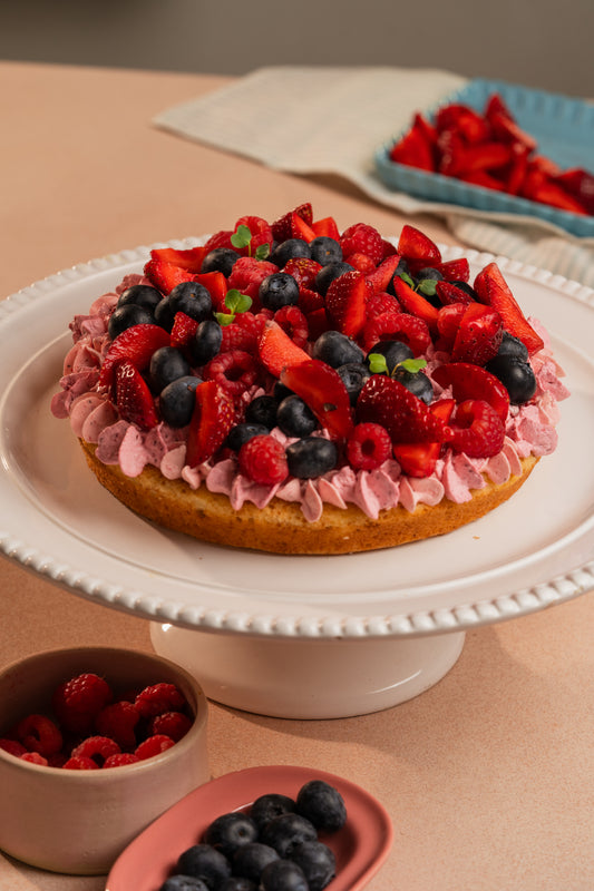 A layered cake topped with fresh strawberries and blueberries, dusted with powdered sugar, presented on a white cake stand with a pink and blue bowl of berries in the foreground.